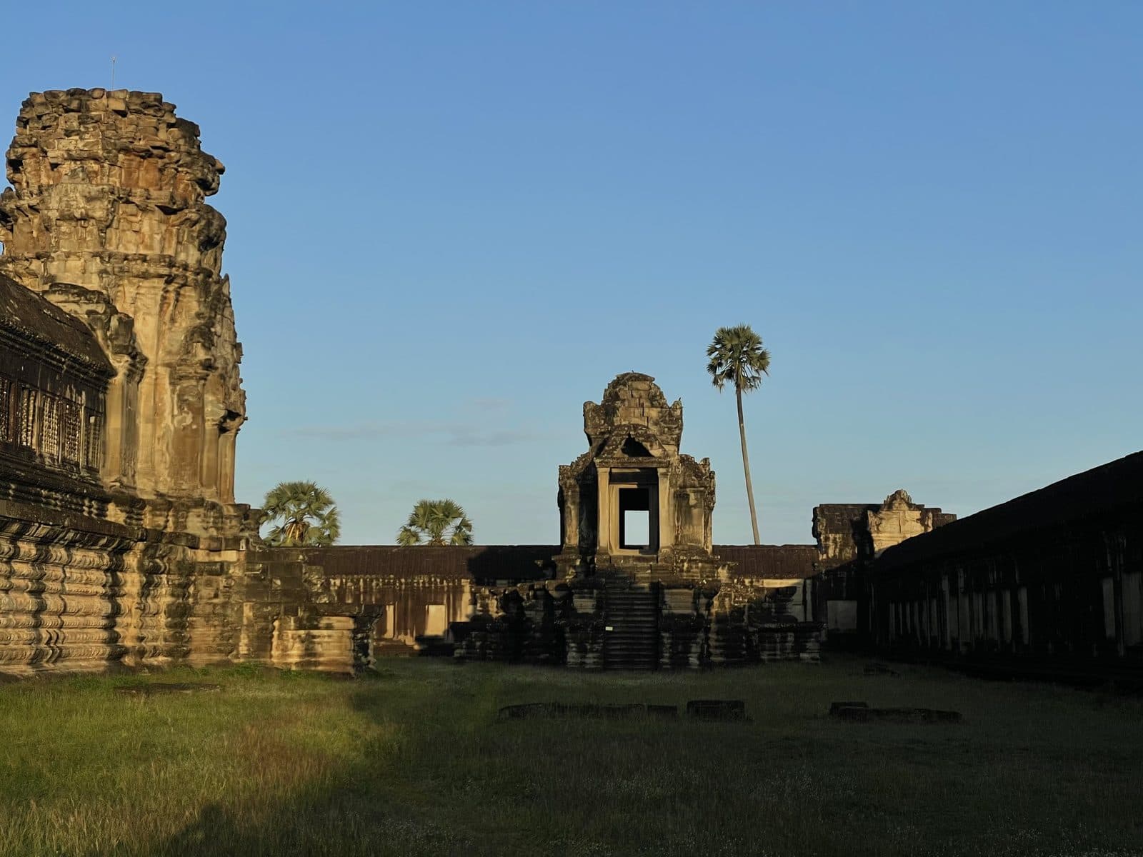 A stone gate and tower of Angkor Wat lit by golden-hour light, with a palm tree behind